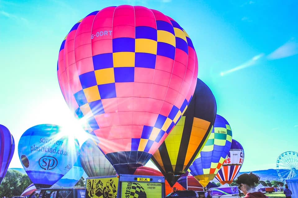 672196495_122181828758606789_6707797523679760710_n - Lydiard Park Colorful hot air balloons ready for dawn ascent at a festival, with bright blue sky and sun glare behind them