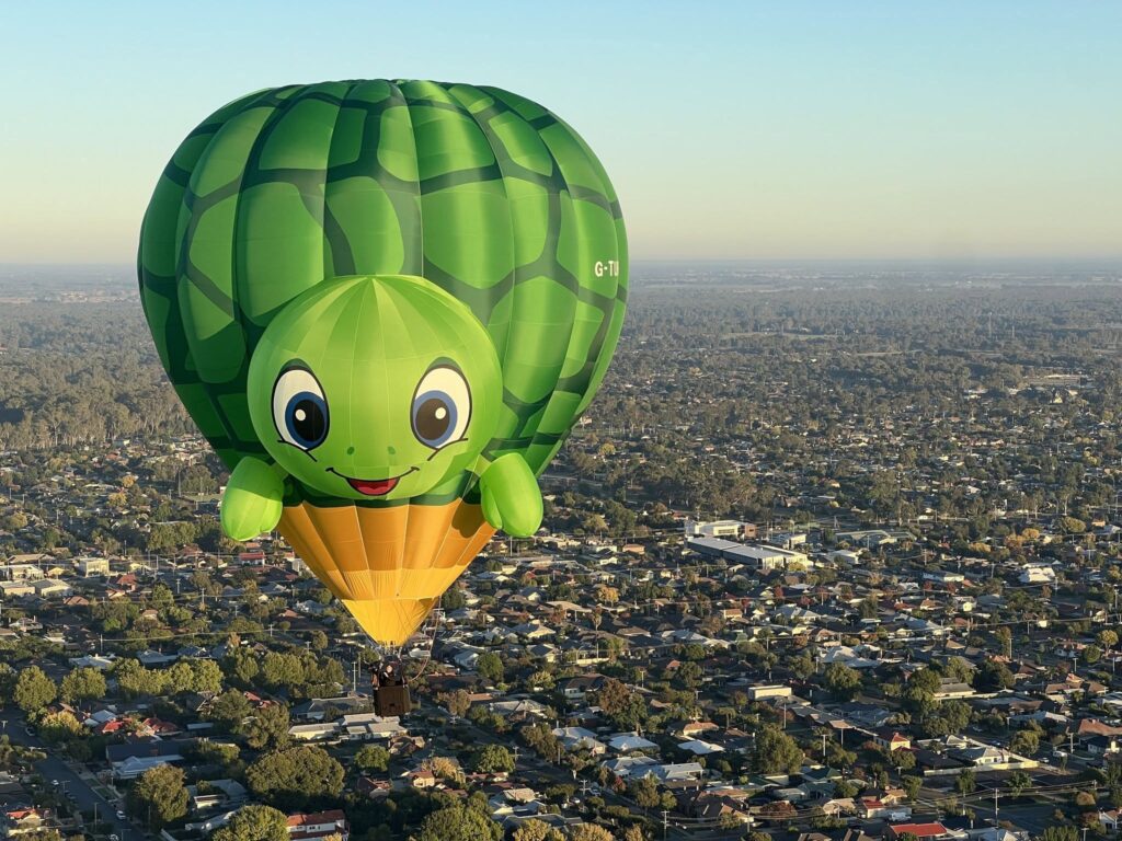 700801009_122184472208606789_216349927385337334_n - Lydiard Park Large green hot air balloon shaped like a friendly turtle with a smiling face, floating over a suburban cityscape.