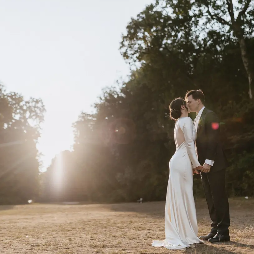 Bride in a white gown and groom in a suit share a kiss outdoors at sunset.