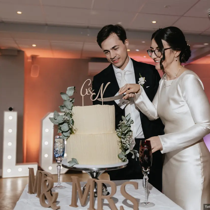 Bride and groom in formal attire cut a cream wedding cake together, with elegant décor and lights in the background.